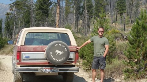 1985 Ford Bronco - A man stands next to the rear of a red 1985 Ford Bronco with a tan top, parked on a dirt road surrounded by pine trees.