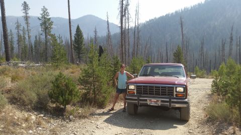 1985 Ford Bronco - A woman stands next to a red 1985 Ford Bronco on a dirt road in a forested area with mountains in the background.