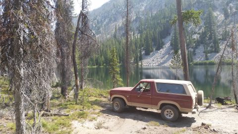 1985 Ford Bronco - A maroon and tan Ford Bronco is parked on a dirt path next to a calm lake, with a steep, tree-covered mountainside in the background.