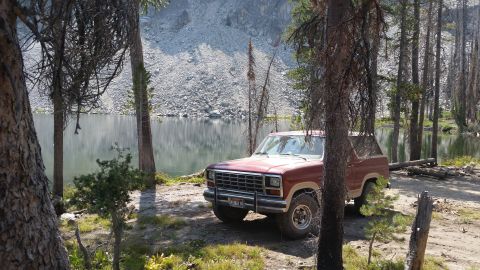 1985 Ford Bronco - A red 1980s Ford Bronco is parked on a dirt path next to a calm lake, with a rocky mountainside in the background and tall pine trees surro…