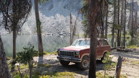1985 Ford Bronco - A red Ford Bronco is parked on a dirt path next to a calm lake, with a rocky mountainside in the background.