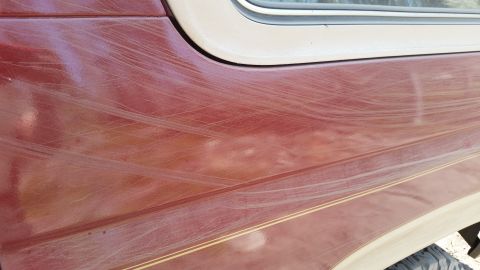 1985 Ford Bronco - A close-up view of the side of a red vehicle, showing numerous light-colored scratches on the paint. A tan trim line runs horizontally acro…