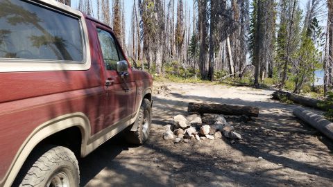 1985 Ford Bronco - The side view of a maroon and tan Ford Bronco parked in a wooded campsite with a fire pit in the foreground.