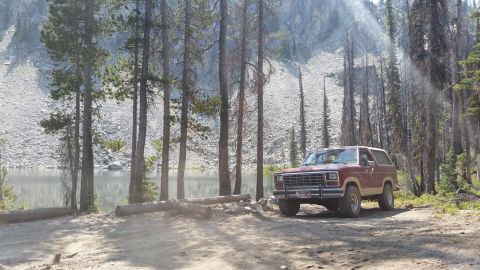 1985 Ford Bronco - A maroon and tan 1980s Ford Bronco is parked on a dirt lot next to a lake with a rocky hillside in the background. Tall pine trees surround…