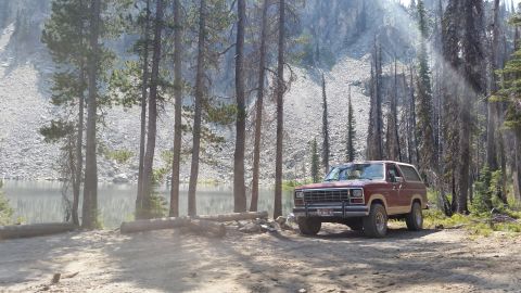 1985 Ford Bronco - A maroon 1980s Ford Bronco is parked on a dirt road next to a lake, surrounded by pine trees and a rocky mountainside. Sunlight streams thr…