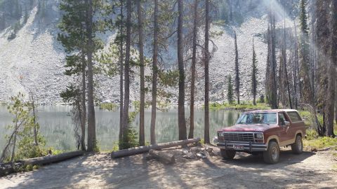 1985 Ford Bronco - A maroon 1985 Ford Bronco is parked on a dirt path next to a calm lake, with a steep, rocky hillside and pine trees in the background.
