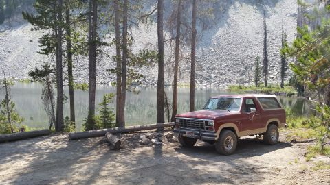 1985 Ford Bronco - A maroon and tan 1980s Ford Bronco is parked on a dirt pull-off next to a calm lake, with a rocky mountainside in the background.
