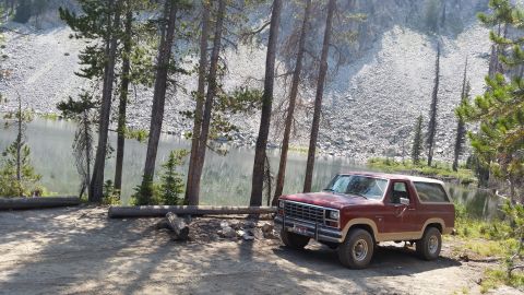 1985 Ford Bronco - A maroon 1980s Ford Bronco is parked on a dirt lot next to a lake with a rocky hillside in the background.