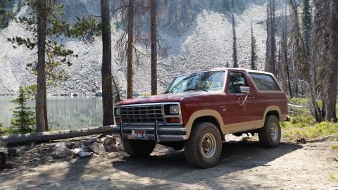 1985 Ford Bronco - A maroon and tan 1985 Ford Bronco is parked on a dirt path next to a calm lake and a rocky hillside. Tall, thin pine trees surround the veh…