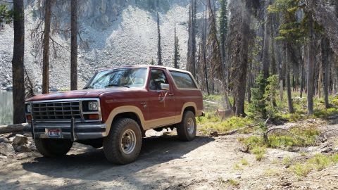 1985 Ford Bronco - A maroon and tan two-door Ford Bronco is parked on a dirt path in a forest with a rocky mountainside and a lake in the background. A deer o…