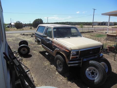 1984 Ford Bronco - A tan and brown 1980s Ford Bronco is parked outdoors on a dirt lot next to a white building. The front of the Bronco is visible, along with…