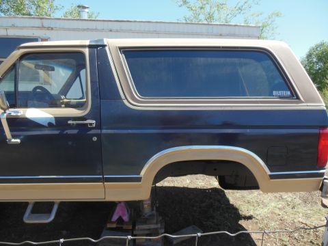 1984 Ford Bronco - The side profile of a dark blue and tan Ford Bronco is shown, lifted on jack stands. A Bilstein sticker is visible on the rear passenger wi…