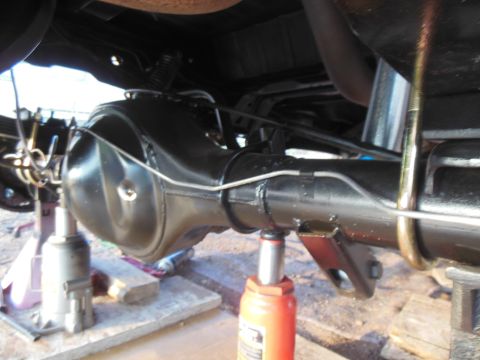 1984 Ford Bronco - The underside of a vehicle shows a black painted rear differential assembly supported by jack stands and a hydraulic jack. Brake lines are…
