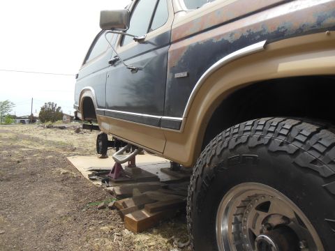 1984 Ford Bronco - A side view of a dark blue and tan Ford Bronco being worked on, lifted on jack stands with a large off-road tire visible. The vehicle shows…