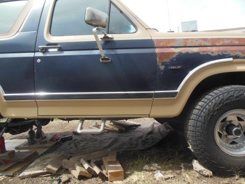 1984 Ford Bronco - The side view of a dark blue and tan Ford Bronco shows rust on the front fender and a chrome trim piece along the lower body. A step bar is…