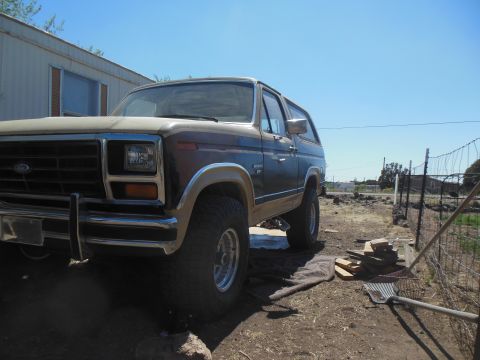 1984 Ford Bronco - A low-angle, front-three-quarter view of a vintage Ford Bronco parked on a dirt lot. The vehicle has a two-tone paint scheme with a tan top…