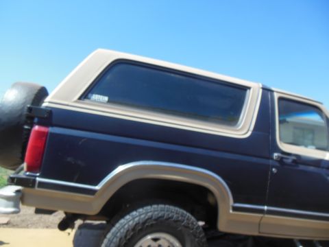 1984 Ford Bronco - The rear passenger side of a dark blue 1980s Ford Bronco with a tan roof and trim is shown against a clear blue sky. A spare tire is mounte…
