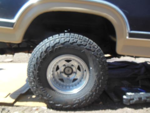 1984 Ford Bronco - A close-up side view of the rear wheel and fender of a dark blue and tan Ford Bronco. The tire is a knobby off-road tire mounted on a chrom…