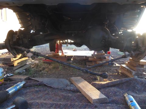 1984 Ford Bronco - The undercarriage of a vehicle, likely an SUV, is supported by wooden blocks and hydraulic jacks. The front axle and steering components ar…
