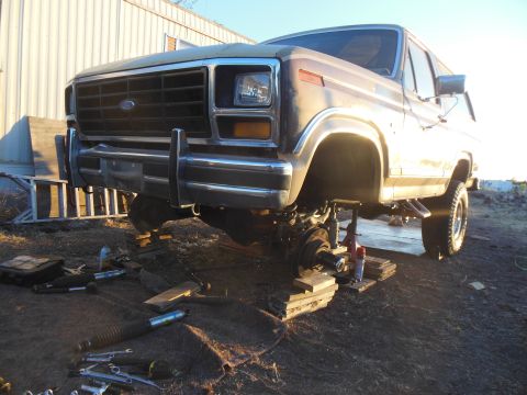 1984 Ford Bronco - A tan 1980s Ford Bronco is jacked up with its front wheel removed, showing suspension components and tools laid out on the ground. The vehi…