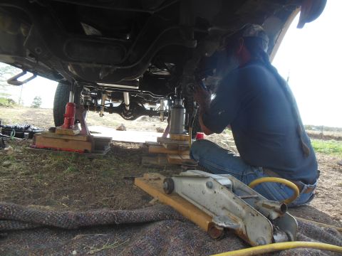 1984 Ford Bronco - A person works on the undercarriage of a vehicle supported by multiple jacks and jack stands outdoors. The vehicle appears to be a truck or…