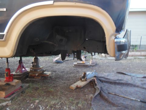 1984 Ford Bronco - The underside of a vehicle, likely a 1980s Ford Bronco, is raised on hydraulic jacks and jack stands in an outdoor setting. The vehicle's f…