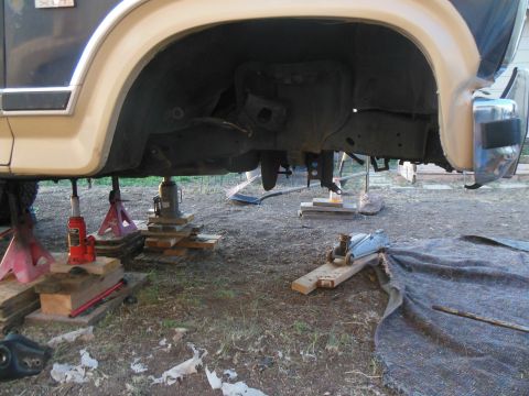 1984 Ford Bronco - The underside of a vehicle is supported by multiple jack stands and wooden blocks. A hydraulic floor jack is positioned on a ramp, ready to…