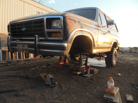 1984 Ford Bronco - A tan 1984 Ford Bronco is raised on jack stands in an outdoor setting with a building in the background. The front of the vehicle is visibl…