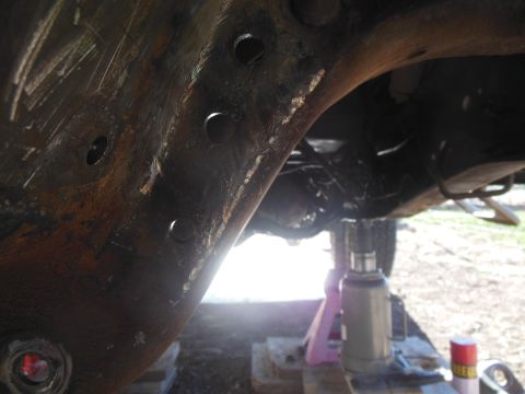 1984 Ford Bronco - Close-up view of the rusty undercarriage of a vehicle, showing a large metal frame with several circular holes. A hydraulic jack and jack s…
