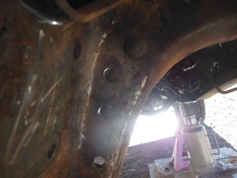 1984 Ford Bronco - The underside of a rusty vehicle frame is shown, with a hydraulic jack and jack stand supporting it. The vehicle appears to be a truck or S…