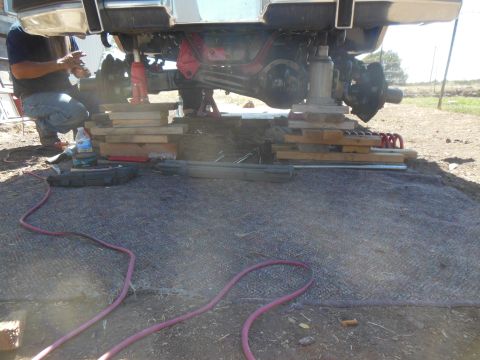 1984 Ford Bronco - A person works on the rear axle of a lifted vehicle, which is supported by wooden blocks and a hydraulic jack. Tools and hoses are visible…