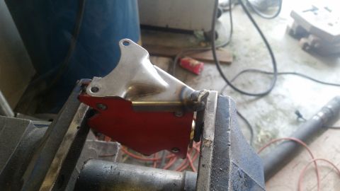 1985 Ford Bronco - A shiny metal bracket with a red plate attached is clamped in a vise. The bracket appears to have been recently welded, with visible weld m…