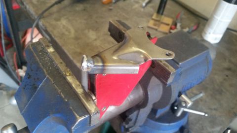 1985 Ford Bronco - A custom metal bracket, likely a vehicle part, is secured in a blue vise on a workbench. The bracket appears to have been recently welded,…