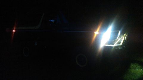1978 Ford Bronco - A dark, nighttime photo shows the front of a dark-colored SUV with its headlights on, illuminating the front end and a grassy area to the r…