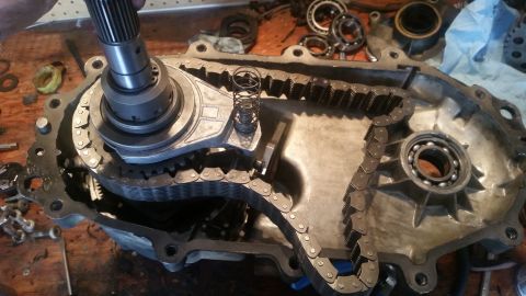 1985 Ford Bronco - A transfer case is disassembled on a wooden workbench, revealing gears and a timing chain. A hand is visible at the top left, holding a sha…