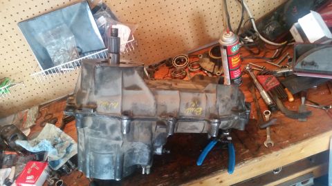 1985 Ford Bronco - A transfer case sits on a workbench with various tools and parts scattered around it. The transfer case is a heavy-duty metal component, li…