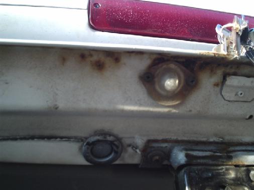 Rusted white rear body panel with drain plugs below a red taillight lens