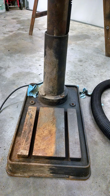 1995 Ford Bronco - The base of a rusty industrial drill press sits on a concrete floor. A black corrugated hose is visible to the right of the drill press.