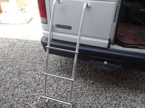 1999 Ford E250 - A silver ladder is attached to the rear of a white Ford E250 van. The van has a black bumper and a hitch receiver visible.