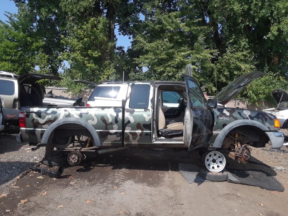 Camouflage Ford Ranger pickup truck in junkyard with doors and hood open, missing wheels.