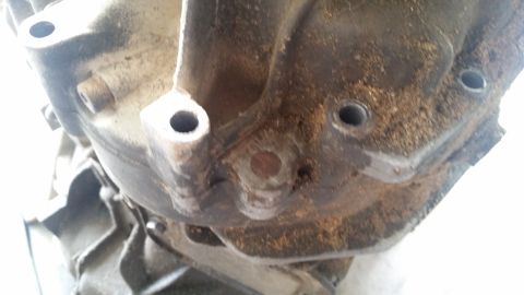 1985 Ford Bronco - A close-up, angled view of a heavily soiled and weathered metal automotive transmission housing with two visible bolt holes and a central b…