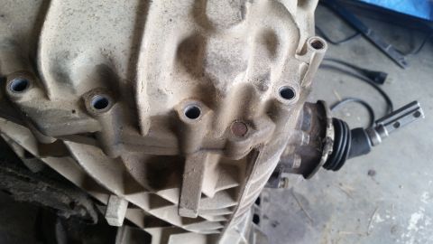 1985 Ford Bronco - A close-up, dusty view of a transmission housing with several bolt holes visible. A rubber boot and part of a shifter mechanism are also vi…