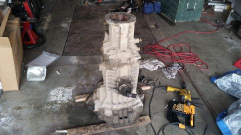 1985 Ford Bronco - A dirty, heavy-duty transmission is laid out on wooden planks on a garage floor. A yellow drill and a box of drill bits are visible to the…