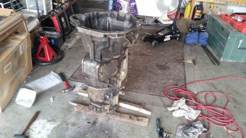 1985 Ford Bronco - A large, heavy-duty transmission casing is propped up on wooden blocks on a garage floor. Various tools and automotive parts are scattered…