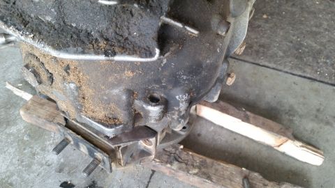 1985 Ford Bronco - A heavily soiled and greasy transmission is supported by wooden blocks and a metal bracket with two threaded bolts. The transmission appear…