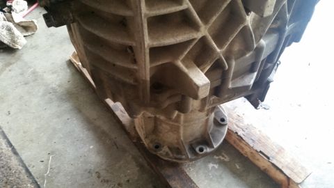 1985 Ford Bronco - A close-up, low-angle view of the underside of a cast-iron transmission housing, resting on wooden planks on a concrete floor. The housing…
