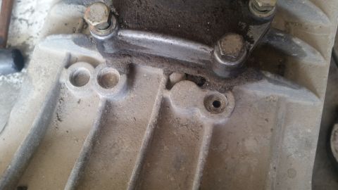1985 Ford Bronco - A close-up view of a dirty, metallic automotive part, likely from a transmission or engine casing, showing bolts and mounting points.