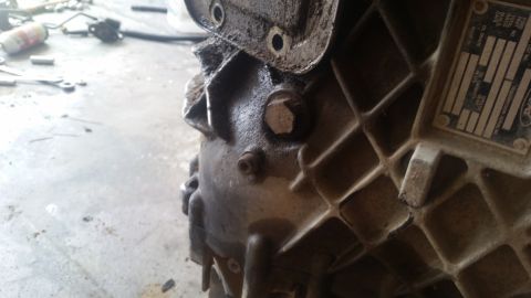 1985 Ford Bronco - A close-up, low-angle shot shows the dirty, cast-iron housing of a vehicle transmission. A metal data plate with Ford branding and specific…