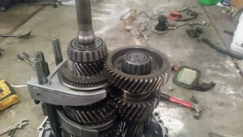 1985 Ford Bronco - A close-up view of a disassembled manual transmission, showing various gears and shafts resting on a concrete floor. Tools like pliers, a h…