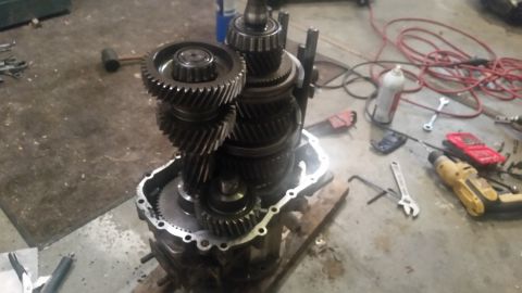 1985 Ford Bronco - A disassembled manual transmission is laid out on a concrete floor, surrounded by various tools including wrenches, a drill, and a mallet.…